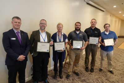 Six people standing in a row looking at the camera. Five are holding Certified Emergency Management Director certificates