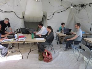 Individuals sitting at tables in a large tent working on computers.