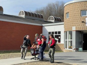 Emergency responders wheeling out a simulated victim from a school on a desk chair. This is simulated and part of an exercise
