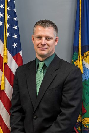 VEM Director Eric Forand posing in front of U.S. and Vermont flags